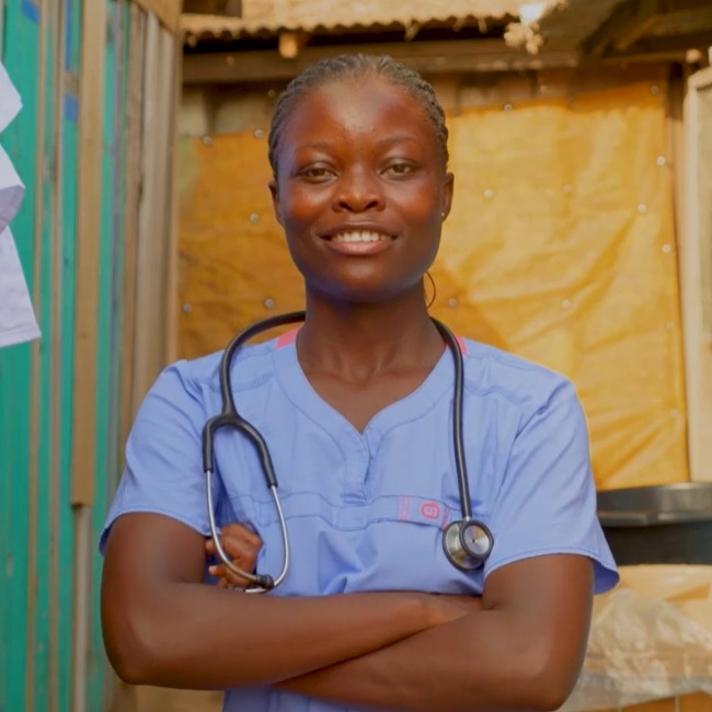 Woman wearing scrubs and a stethoscope around her shoulders smiles in to the camera