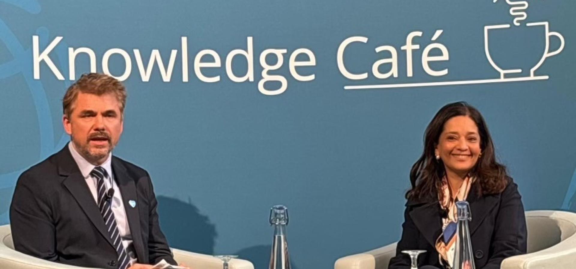 Priya Basu and Scott Pendergast sit in front of a backdrop which reads Knowledge Café, with a lightbulb over a mug