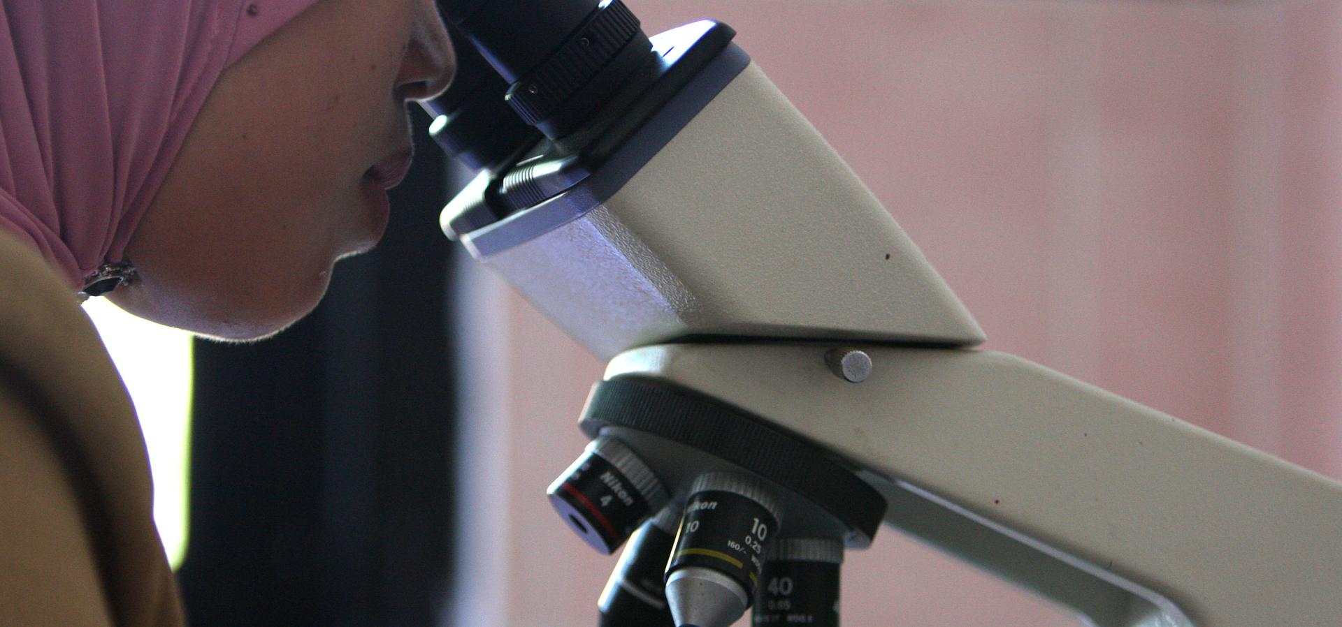 Indonesian woman wearing a headscarf looks into a microscope