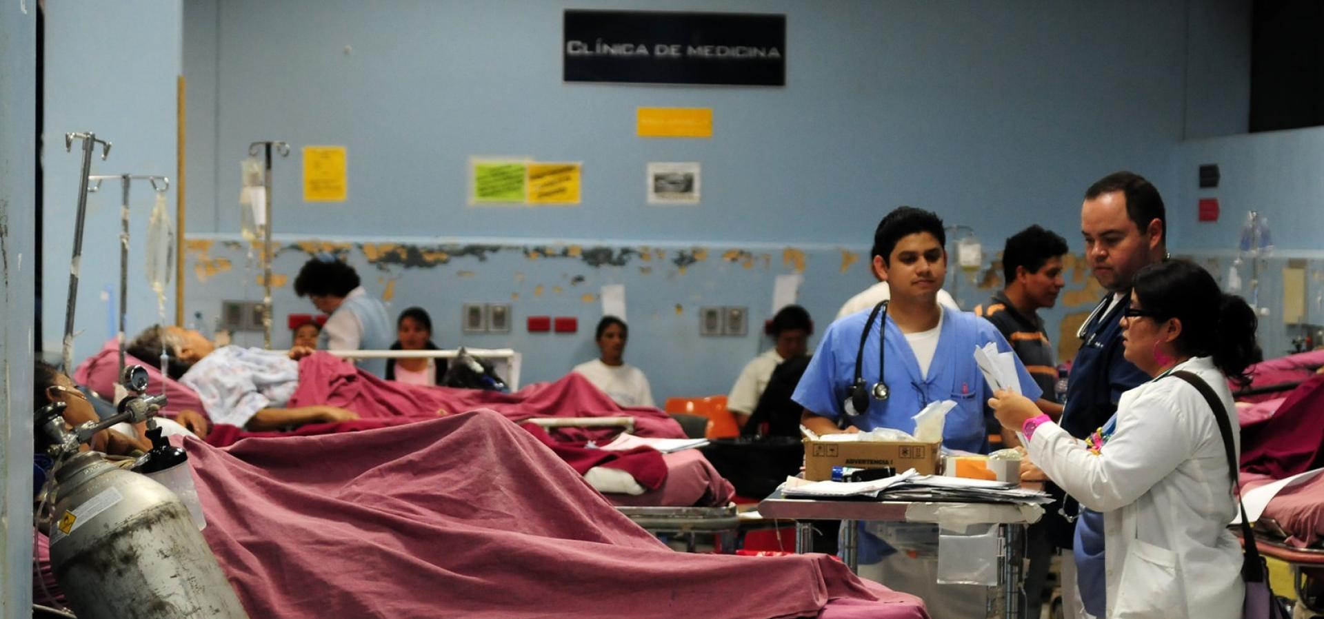 Health workers look over paperwork while patients lie in hospital beds. The words Clinica de medicina are written on the wall behind them