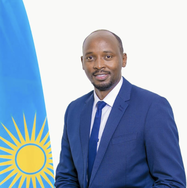 African man wearing a blue blazer looks into camera, with the flag of Rwanda off his right shoulder