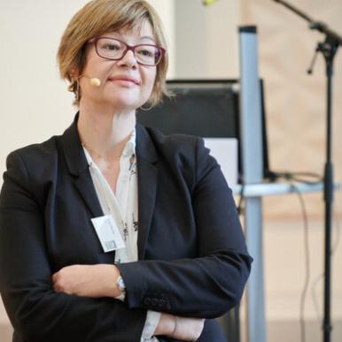 Woman in a black blazer, wearing glasses, stands crossing her arms and wearing a microphone