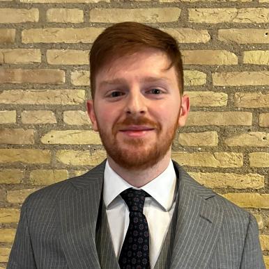 Man wearing a gray suit stands in front of a brick wall