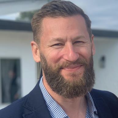 Man with a light beard wearing a blue suit, stands outside in front of a house and looks into the camera