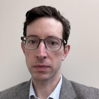 White man with short brown hair, wearing glasses and a gray suit, looks into the camera