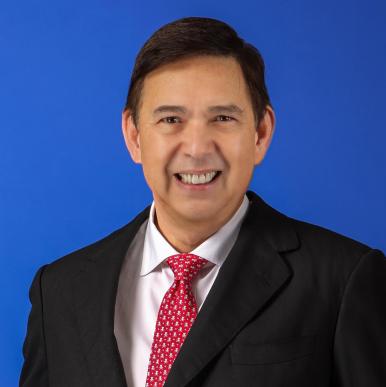 Man wearing a black suit with a red tie, stands in front of a bright blue background and smiles towards the camera