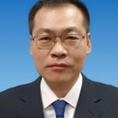 Man in black suit wearing glasses looks into the camera, in front of a blue background