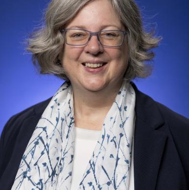 White woman wearing blue glasses, a white scarf with blue lines, stand in front of a blue backdrop and looks into the camera