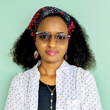 Woman with dark curly hair looks into the camera, wearing glasses, a colorful headband, and dotted shirt