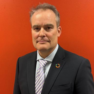 Man wearing a black suit with an SDG pin on the lapel stands in front of a burnt-orange background and looks into the camera