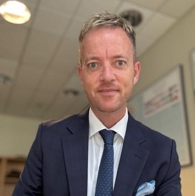 Man wearing a blue suit with a white shirt and blue tie smiles into the camera, while sitting in an office