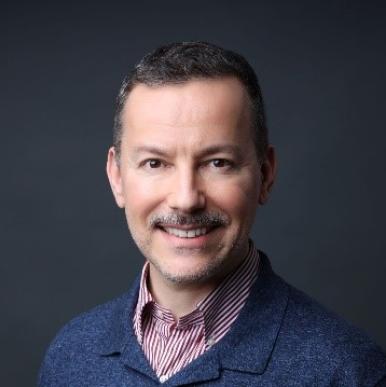 Man with a light moustache smiles into the camera, wearing red shirt and blue jacket