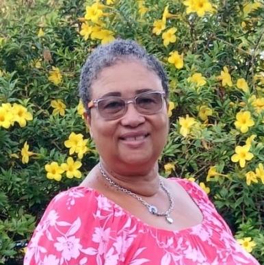 Woman with short hair, wearing a pink dress and glasses, smiles into the camera. She stands in front of a bush with bright yellow flowers