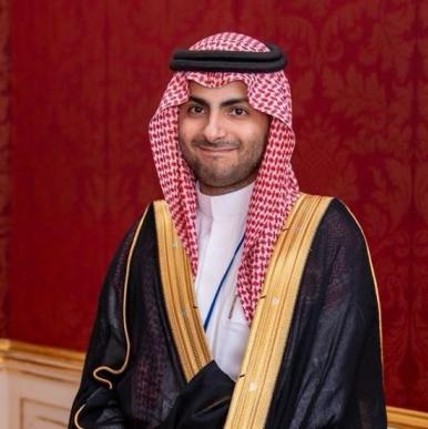 Saud M AlBarrak headshot Man wearing a red headscarf and black robe with gold trim, smiles into the camera