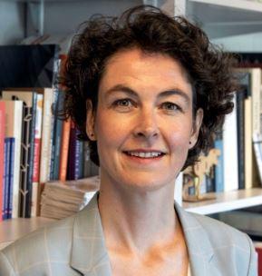 Caucasian woman with short hair in a grey blazer, stnads in front of a bookshelf and looks into the camera