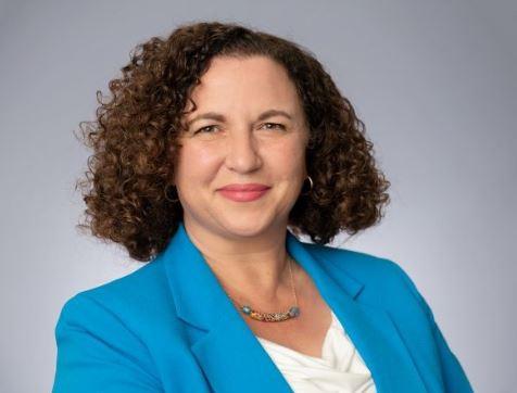 Woman with short, brown curly hair wearing a bright, light blue blazer, looks into the camera