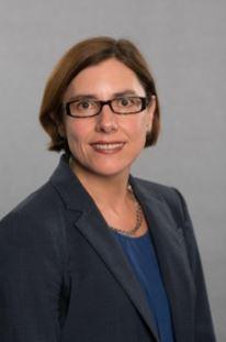 Caucasian woman with short brown hair, wearing glasses and a black blazer, looks into the camera