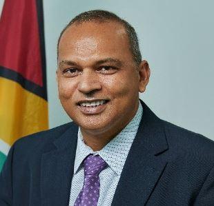 Man in a blue suit with a purple tie sits in front of the flag of Guyana