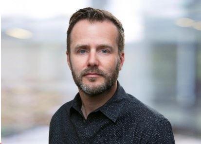 Causian man with a short beard, wearing a blue collared shirt, looks into the camera