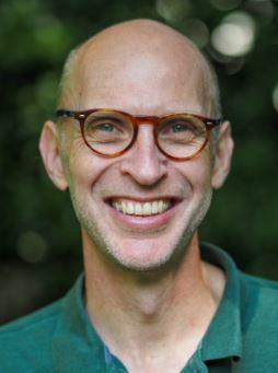 Caucasian man without hair, wearing red glasses and a green polo shirt, smiles into the camera