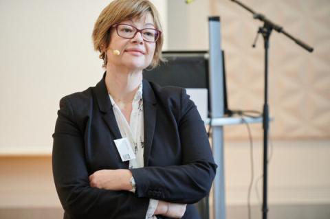 Woman in a black blazer, wearing glasses, stands crossing her arms and wearing a microphone