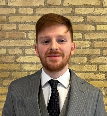 Man wearing a gray suit stands in front of a brick wall