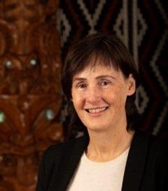 Woman with brown hair with bangs, wearing a white shirt and black blazer, stands and smiles into the camera