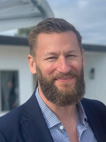 Man with a light beard wearing a blue suit, stands outside in front of a house and looks into the camera