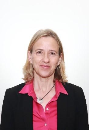 Woman with blonde hair, wearing a pink blouse and black blazer, stands in front of a white background 