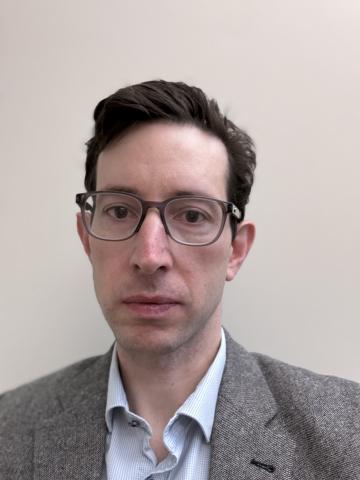 White man with short brown hair, wearing glasses and a gray suit, looks into the camera