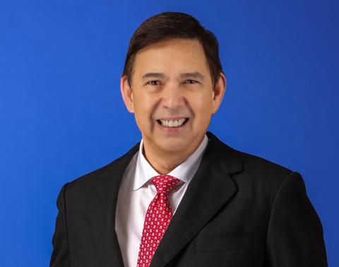 Man wearing a black suit with a red tie, stands in front of a bright blue background and smiles towards the camera