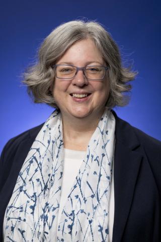 White woman wearing blue glasses, a white scarf with blue lines, stand in front of a blue backdrop and looks into the camera