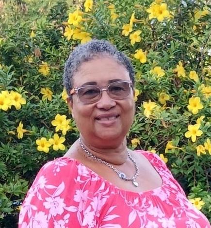 Woman with short hair, wearing a pink dress and glasses, smiles into the camera. She stands in front of a bush with bright yellow flowers