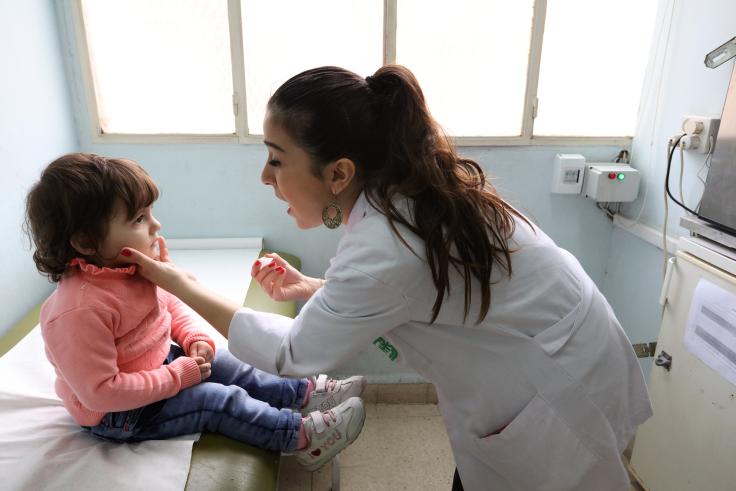 Nurse wearing a white bends down to a child and makes the motion to open their mouth, with a liquid dropper in one hand