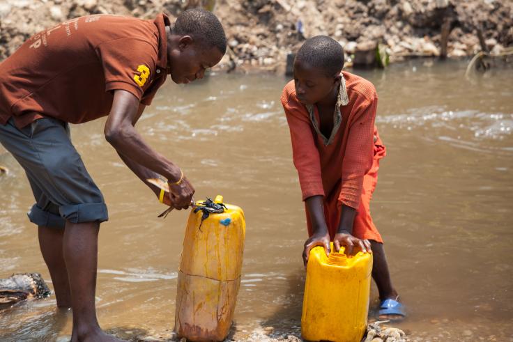 Two boys stand in a muddy river, gathering water into yellow jugs